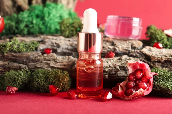 Cosmetic dropper bottle with green moss, pomegranate and tree bark on red background, closeup