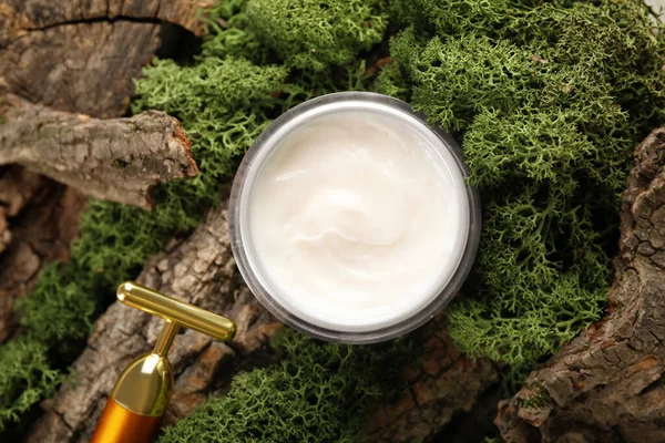 Jar of cream with facial massage tool and green moss on tree bark, closeup