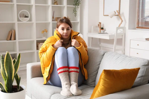 Frozen young woman in down jacket sitting on sofa at home