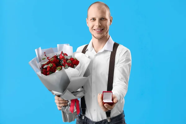 Young man with bouquet of flowers and engagement ring on blue background. Valentine's Day celebration