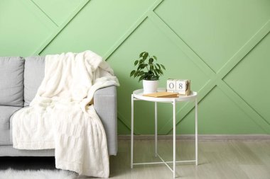 Table with houseplant, book, cube calendar and grey sofa near green wall