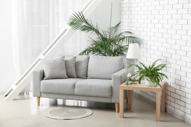 Table with lamp, houseplants and grey sofa in light living room