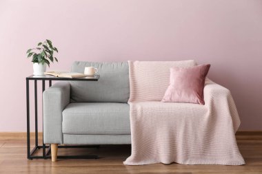 Table with houseplant, cup, book and grey sofa near pink wall