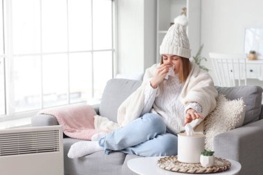 Ill young woman in winter clothes warming near radiator at home