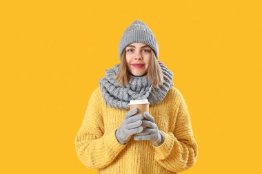 Frozen young woman in winter clothes with cup of coffee on yellow background