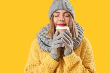 Frozen young woman in winter clothes with cup of coffee on yellow background