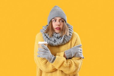 Frozen young woman in winter clothes with cup of coffee on yellow background