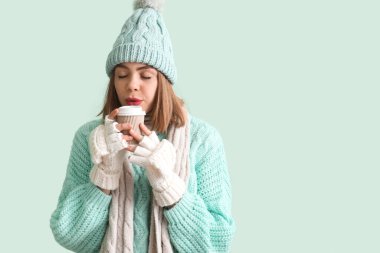 Frozen young woman in winter clothes with cup of hot coffee on green background