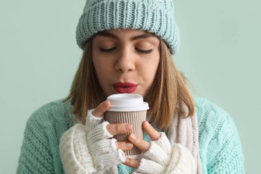 Frozen young woman in winter clothes with cup of hot coffee on green background, closeup
