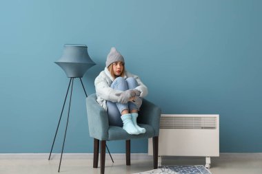 Frozen young woman in winter clothes heating near radiator at home