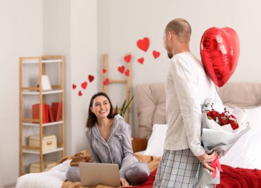 Young man surprising his wife with flowers and balloon in bedroom on Valentine's Day