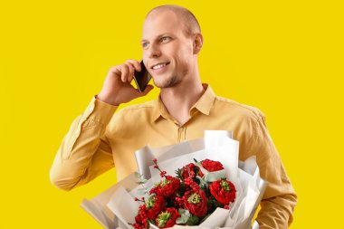 Young man with bouquet of flowers talking by mobile phone on yellow background. Valentine's Day celebration