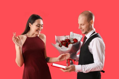 Young man greeting his wife with bouquet of flowers and gift on red background. Valentine's Day celebration