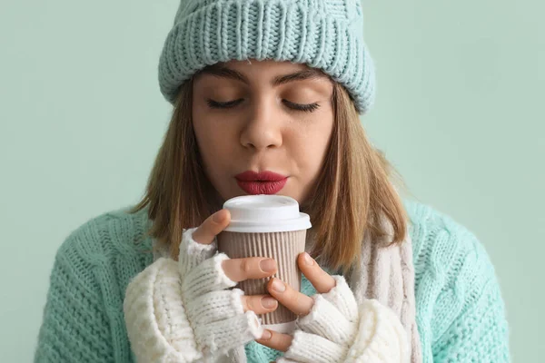 Frozen young woman in winter clothes with cup of hot coffee on green background, closeup
