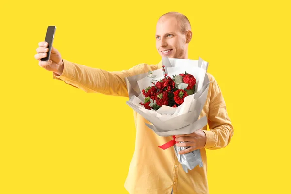 Young man with bouquet of flowers taking selfie on yellow background. Valentine's Day celebration