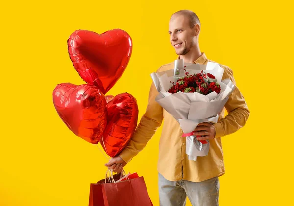 Young man with bouquet of flowers, bags and balloons on yellow background. Valentine\'s Day celebration