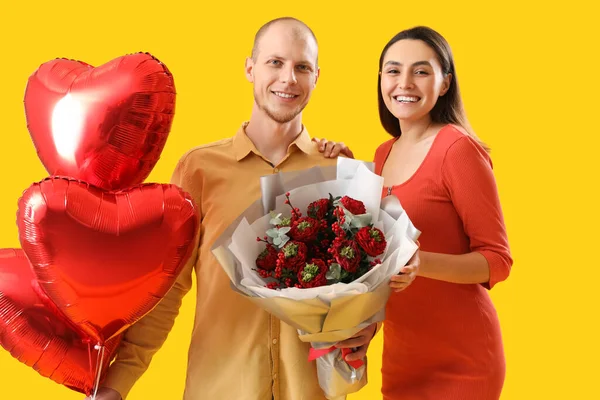 Young couple with bouquet of flowers and balloons on yellow background. Valentine\'s Day celebration