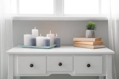 Burning candles, books and houseplant on table near window