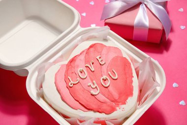 Plastic lunch box with heart-shaped bento cake and gift on pink background. Valentine's Day celebration