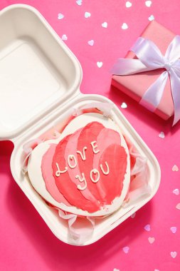 Plastic lunch box with heart-shaped bento cake and gift on pink background. Valentine's Day celebration