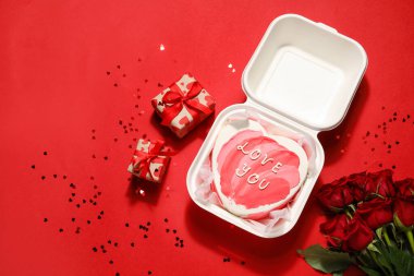 Plastic lunch box with heart-shaped bento cake, gifts and rose flowers on red background. Valentine's Day celebration