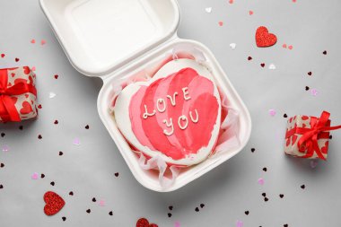 Plastic lunch box with heart-shaped bento cake and gifts on grey background. Valentine's Day celebration