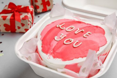 Plastic lunch box with heart-shaped bento cake and gifts on grey background, closeup. Valentine's Day celebration