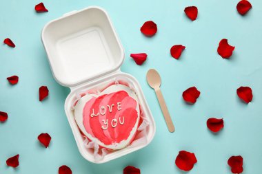 Plastic lunch box with heart-shaped bento cake, spoon and rose petals on turquoise background. Valentine's Day celebration