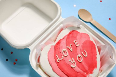 Plastic lunch box with heart-shaped bento cake and spoon on blue background, closeup. Valentine's Day celebration