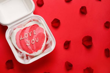Plastic lunch box with heart-shaped bento cake and rose petals on red background. Valentine's Day celebration