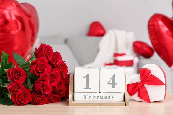 Calendar with date 14 FEBRUARY, gift and roses on table in living room, closeup