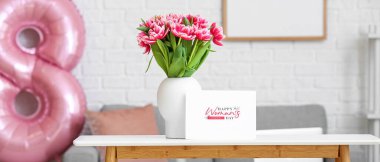 Vase with pink tulips and greeting card with text HAPPY WOMAN'S DAY on table in room