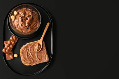 Board with bowl of tasty hazelnut butter, toast and chocolate on dark background