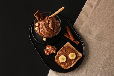 Board with bowl of tasty hazelnut butter, toast, chocolate and cinnamon on dark background