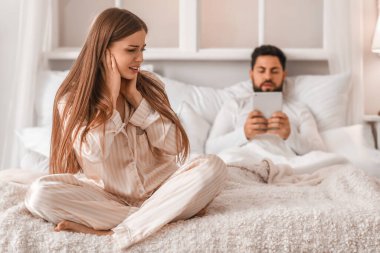 Young woman cannot sleep because of her husband using tablet computer in bedroom