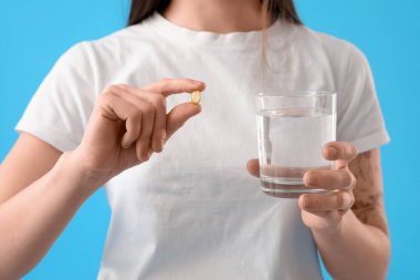 Young woman with glass of water taking vitamin supplement on blue background, closeup