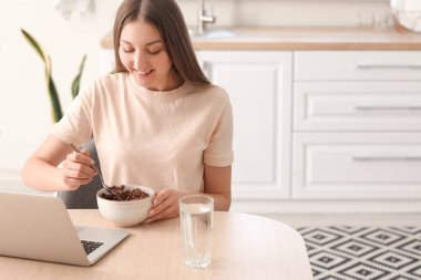 Beautiful woman with laptop eating corn balls at table in kitchen