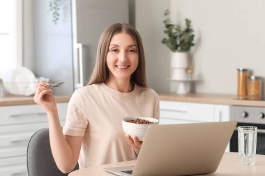 Beautiful woman with laptop eating corn balls at table in kitchen