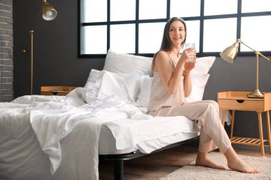 Beautiful woman with glass of water sitting on bed at home
