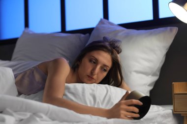 Sleepy young woman with alarm clock lying in bed at night