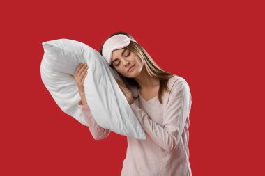 Beautiful woman with sleeping mask and pillow on red background