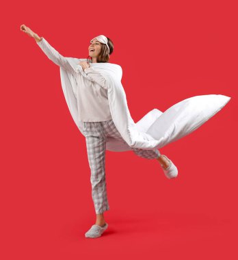 Beautiful woman with sleeping mask and blanket jumping on red background
