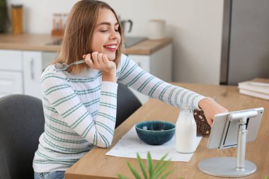Young woman with spoon using tablet computer at table in kitchen