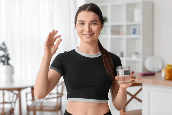 Sporty young woman with vitamin supplement and glass of water at home