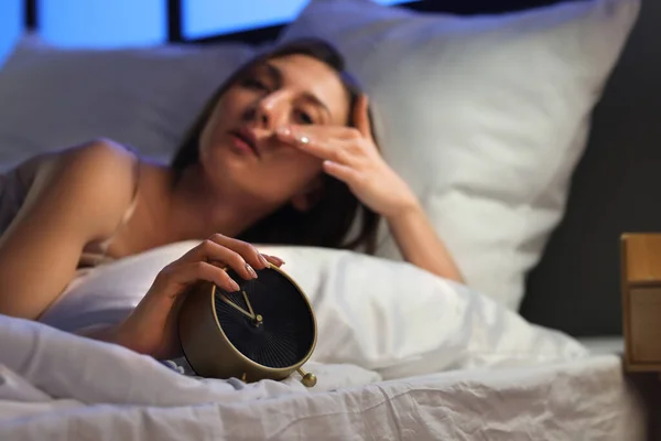 Sleepy young woman with alarm clock lying in bed at night, closeup