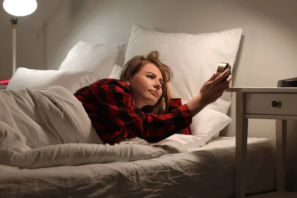 Upset young woman with alarm clock lying in bed at night