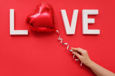 Female hand with word LOVE made of white letters and foil balloon on red background. Valentine's Day celebration