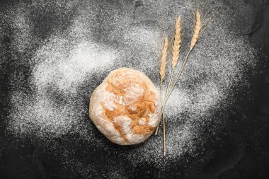 Loaf of fresh bread, wheat ears and flour on black table