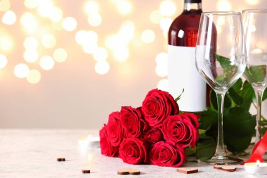 Bottle of wine, rose flowers and glasses on white table against blurred lights. Valentine's Day celebration