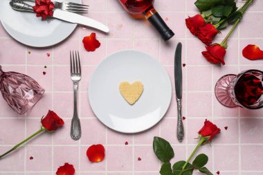 Table setting with bottle of wine, rose flowers, petals and glasses on pink tile background. Valentine's Day celebration
